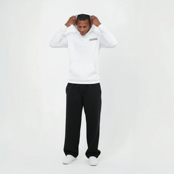 Sweat à capuche blanc sans cordon pour homme, modèle "Stand Clair" de Tourismo Clothing. Hoodie coupe droite avec poche kangourou, porté avec un pantalon noir ample et des baskets blanches. Logo brodé noir "TOURISMO" sur la poitrine côté cœur, accompagné d’un motif damier discret. Le mannequin est debout en studio, en train d’ajuster sa capuche, tête légèrement baissée. Fond blanc épuré, éclairage homogène. Look streetwear inspiré de l’univers de la course automobile, entre technicité et style minimaliste.