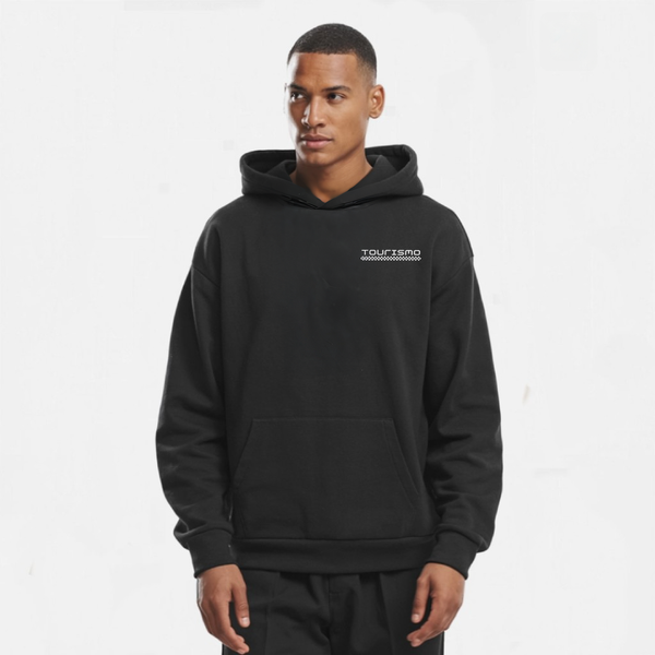 Sweat à capuche noir sans cordon pour homme, modèle "Vue Latérale" de Tourismo Clothing. Hoodie coupe droite avec capuche doublée, poche kangourou et logo brodé blanc "TOURISMO" accompagné d’un motif damier sur la poitrine côté gauche. Le mannequin est en position 3/4, regard tourné vers la droite, bras détendus. Fond blanc studio, éclairage neutre qui souligne la structure du tissu. Tenue streetwear inspirée par le design automobile, alliant sobriété, performance et identité visuelle forte.