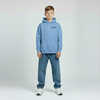 Enfant portant le hoodie bleu Riviera Charles Leclerc, coupe ample, logo poitrine visible, fond studio gris, jambes légèrement écartées.